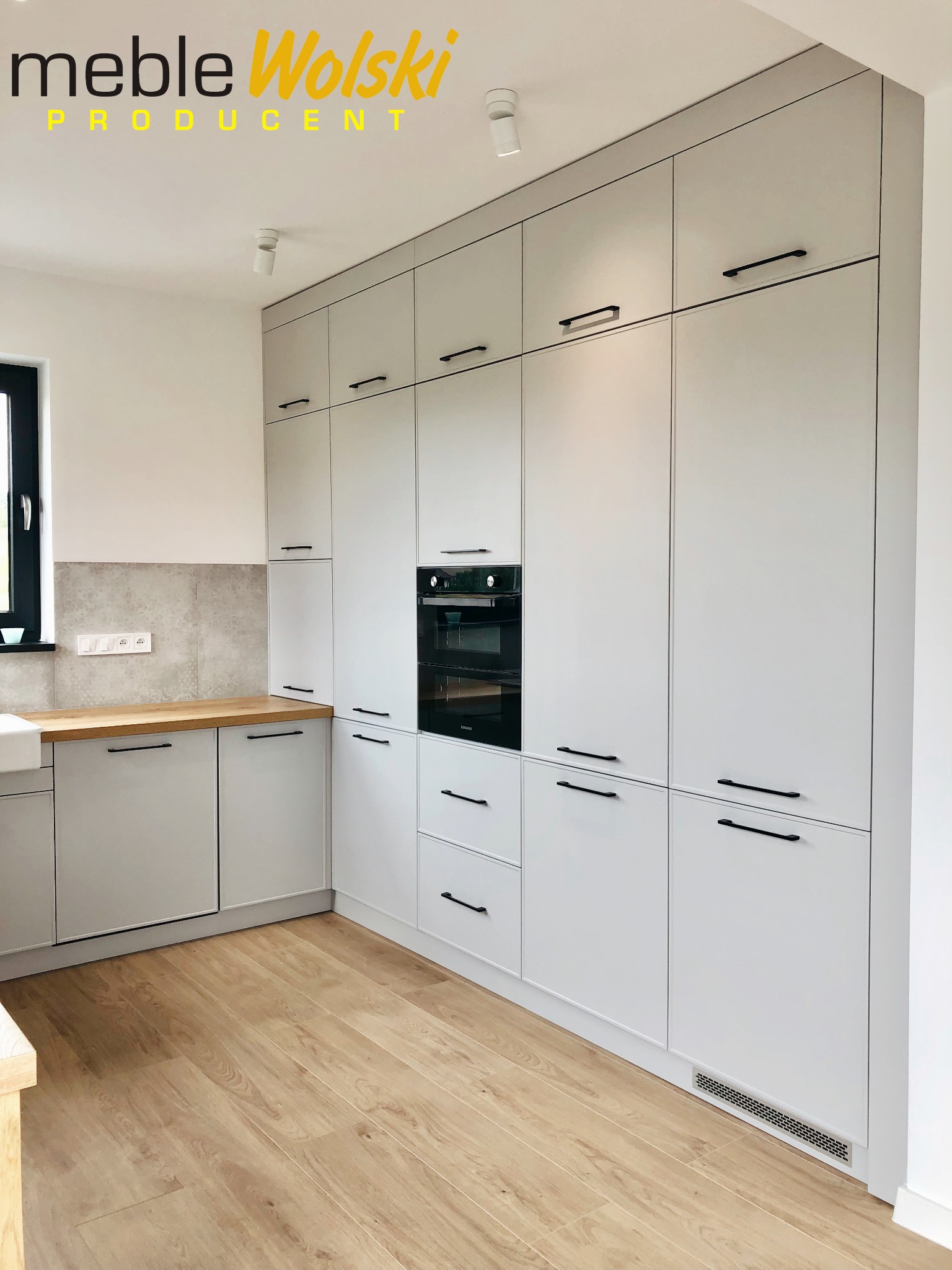 Jasnoszara zabudowa kuchenna z czarnymi uchwytami, wbudowanym piekarnikiem i drewnianym blatem, uzupełniona o logo producenta w górnym rogu.