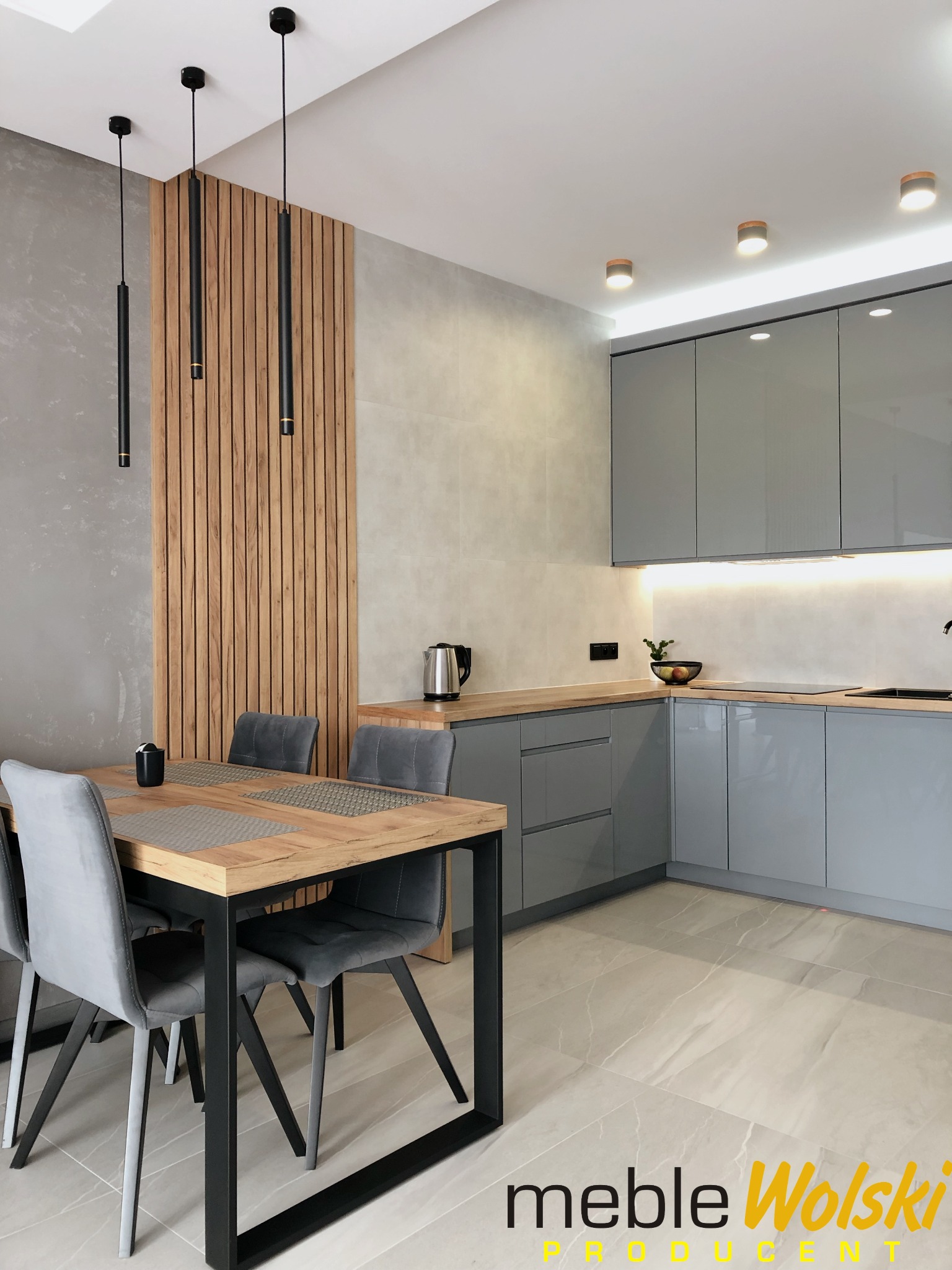 Nowoczesna kuchnia z szarymi frontami i drewnianym blatem, stół z metalowymi nogami i szarymi krzesłami, drewniana ścianka działowa z oświetleniem, minimalistyczne lampy wiszące.
