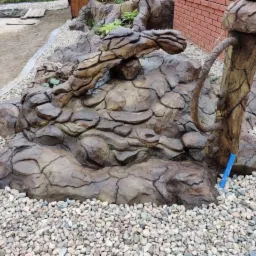 Ozdobny skalniak imitujący naturalny krajobraz z elementami drewna, otoczony drobnymi kamieniami w Olszynie, z widocznym fragmentem ceglanego budynku w tle.