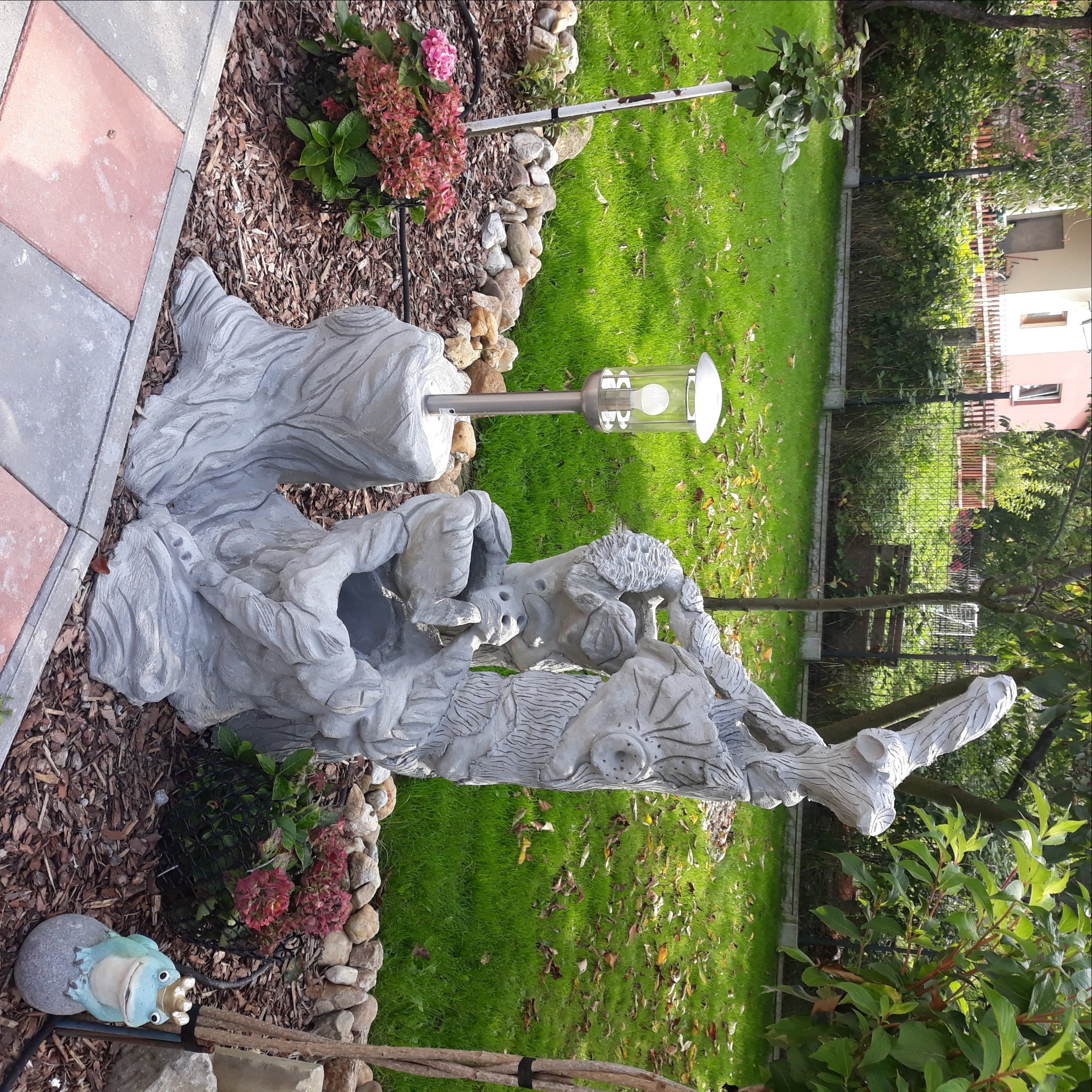 Ozdobna betonowa rzeźba w kształcie pnia drzewa z wbudowaną lampą ogrodową, otoczona korą, kamieniami i roślinami, na tle trawnika.