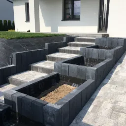 Schody zewnętrzne z kostki brukowej prowadzące do domu, z wydzielonymi miejscami na rośliny, wyłożonymi czarną folią. Część z nich wypełniona ziemią, na pierwszym planie ułożona kostka brukowa.