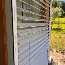 Zamknięte, białe żaluzje fasadowe na tle zielonego ogrodu i błękitnego nieba, widoczne z bliska, z fragmentem drewnianej elewacji budynku.