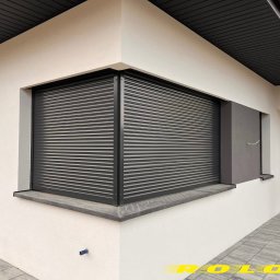 ROLO - Producent Rolet Zewnętrznych Sebastian Włodek - Zamknięta, grafitowa roleta zewnętrzna na oknie narożnym budynku o jasnej elewacji. Widoczny fragment dachu i chodnika z szarych płyt.