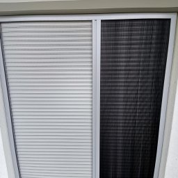 ROLO - Producent Rolet Zewnętrznych Sebastian Włodek - Szara, pozioma roleta zwinięta obok czarnej moskitiery plisowanej w szarej ramie, zamontowane w jasnym otworze okiennym. Widoczne logo firmy.