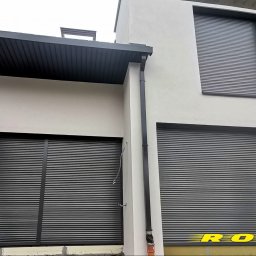 ROLO - Producent Rolet Zewnętrznych Sebastian Włodek - Elewacja budynku z trzema oknami zabezpieczonymi szarymi roletami zewnętrznymi. Widoczne orynnowanie i fragment dachu. Na dole żółta izolacja.