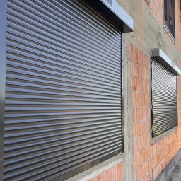 ROLO - Producent Rolet Zewnętrznych Sebastian Włodek - Zamknięta, grafitowa roleta zewnętrzna na tle surowej ściany z cegły i betonu. Widoczne okno z zamontowaną roletą. Ujęcie z dołu, podkreślające solidność konstrukcji.