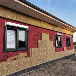 Dom w trakcie ocieplania, widoczne fragmenty elewacji z zamontowanymi oknami i częściowo nałożoną izolacją termiczną, przygotowaną do dalszych prac wykończeniowych.