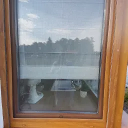 Drewniane okno z zamontowaną moskitierą, widok na zewnątrz z lekkim odbiciem wnętrza, w tle mikser kuchenny i roślina doniczkowa.