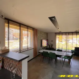 Jasne drewniane żaluzje poziome w nowoczesnym salonie z szarymi płytkami podłogowymi i zielonymi krzesłami przy białym stole. Widoczne elementy dekoracyjne na blacie.