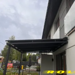 Nowoczesna, czarna pergola tarasowa z płaskim dachem, przylegająca do jasnego budynku z szklaną balustradą balkonową, widok na ogród z elementami placu zabaw.