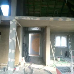 Surowy stan poddasza w trakcie prac wykończeniowych: widoczne elementy konstrukcyjne, częściowo wykończone ściany z widocznymi plamami po szpachlowaniu, rozłożone narzędzia i przewody na podłodze...