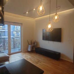 DamBud ( ͡° ͜ʖ ͡°) - Jasne wnętrze salonu z drewnianą podłogą i balkonem. Widoczne lampy wiszące, telewizor na ścianie, sofa i krzesła zabezpieczone folią. Nowoczesny design.