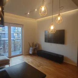 Jasne wnętrze salonu z drewnianą podłogą i balkonem. Widoczne lampy wiszące, telewizor na ścianie, sofa i krzesła zabezpieczone folią. Nowoczesny design.