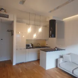 Nowoczesne wnętrze: parkiet, beżowa kuchnia z wyspą, sofa i minimalistyczne obrazy. Oświetlenie punktowe i wiszące lampy dodają elegancji.