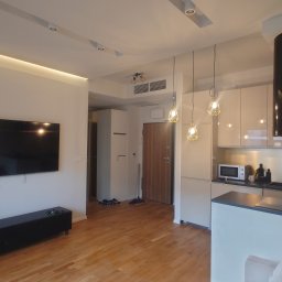 DamBud ( ͡° ͜ʖ ͡°) - Jasne wnętrze mieszkania z drewnianą podłogą, widok na salon z telewizorem i otwartą kuchnią z nowoczesnym okapem oraz designerskimi lampami wiszącymi.