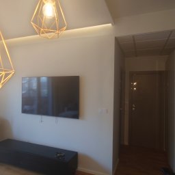 DamBud ( ͡° ͜ʖ ͡°) - Jasne wnętrze po odświeżeniu: minimalistyczny salon z zawieszonym telewizorem, nowoczesnymi lampami i krzesłami zabezpieczonymi folią ochronną.