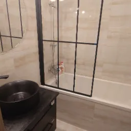 Nowoczesna łazienka z czarną umywalką nablatową, kabiną prysznicową typu walk-in z czarnymi szprosami i beżowymi płytkami imitującymi marmur.