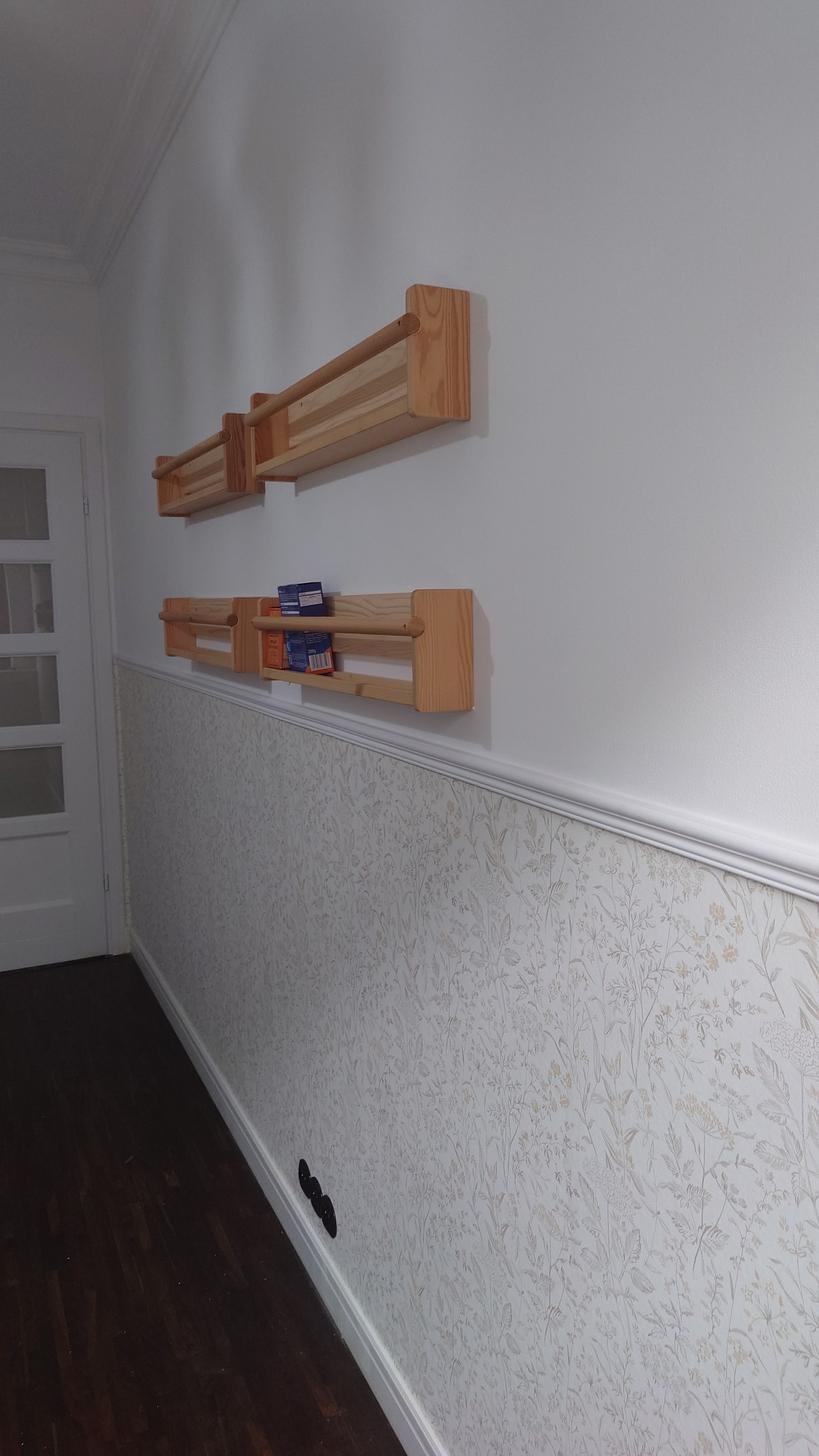 Ściana w przedpokoju z tapetą w kwiaty i dwiema drewnianymi półkami, na jednej z nich stoją pudełka.