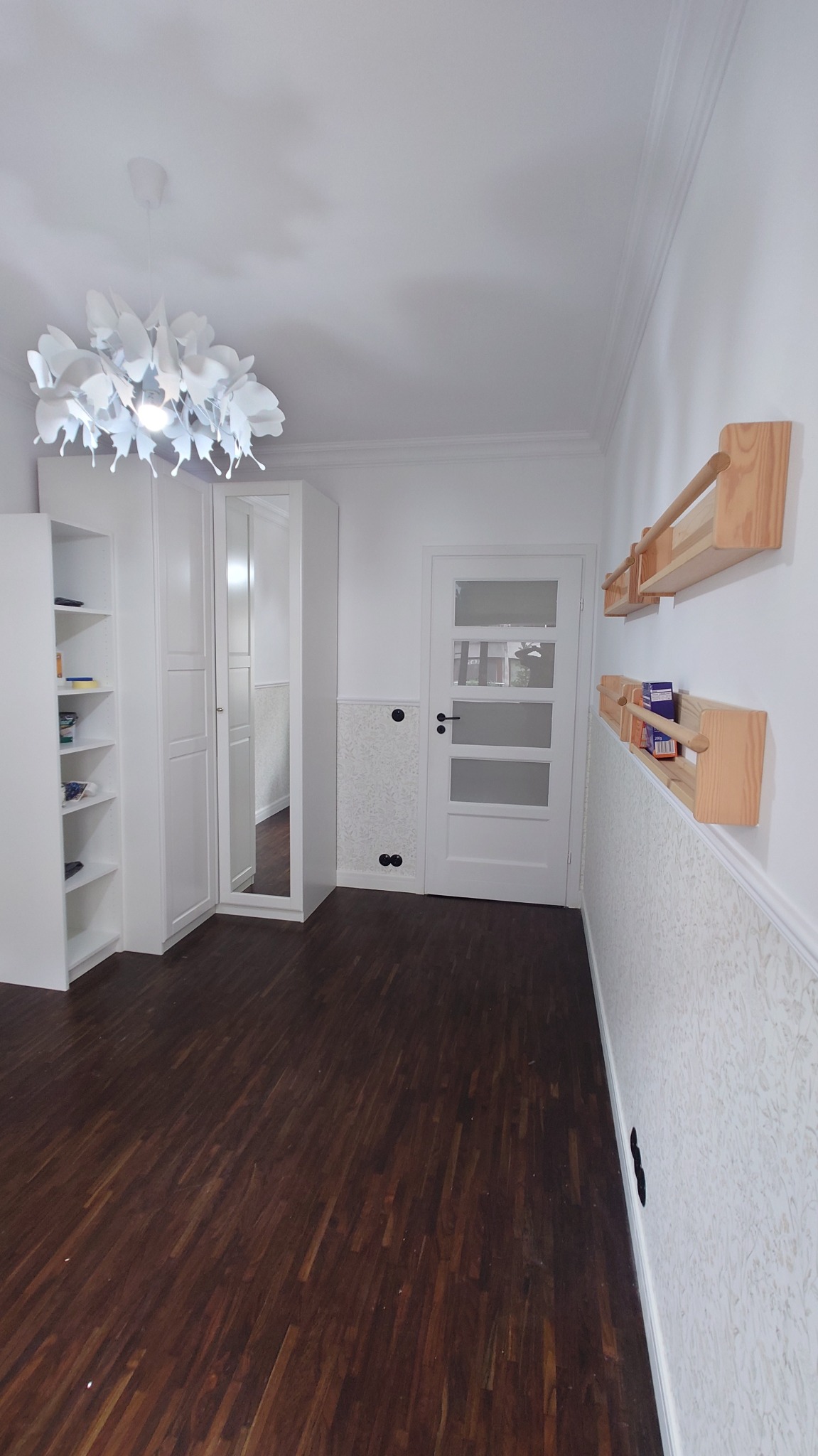 Jasne wnętrze pokoju po remoncie z ciemną podłogą, białymi meblami, ozdobną lampą i półkami na ścianie.