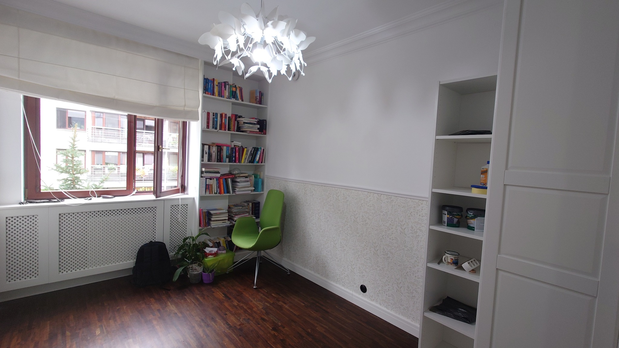 Jasne wnętrze pokoju po remoncie: parkiet w ciemnym odcieniu drewna, białe ściany z dekoracyjną tapetą w dolnej części, białe meble w zabudowie, okno z żaluzjami rzymskimi, designerska lampa...