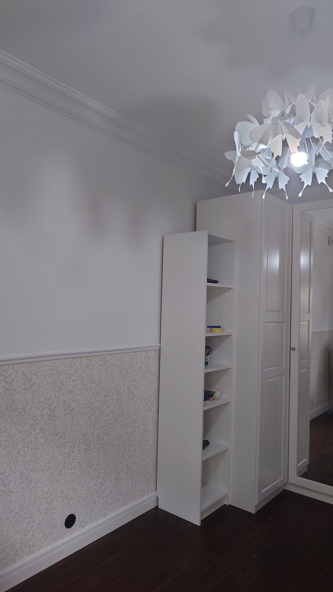 Pokój po remoncie: ściana z tapetą w kwiaty, białe meble systemowe z półkami i szafką, ozdobny żyrandol w kształcie motyli, ciemna podłoga.