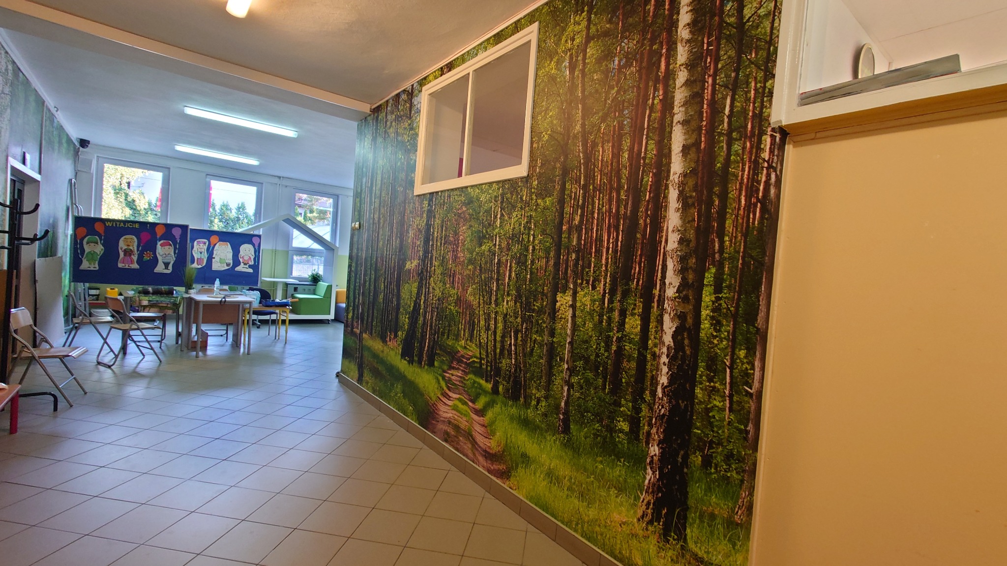 Korytarz szkolny z fototapetą leśną na ścianie, biała rama okienna na fototapecie, w tle klasa z dekoracjami i tablicami.