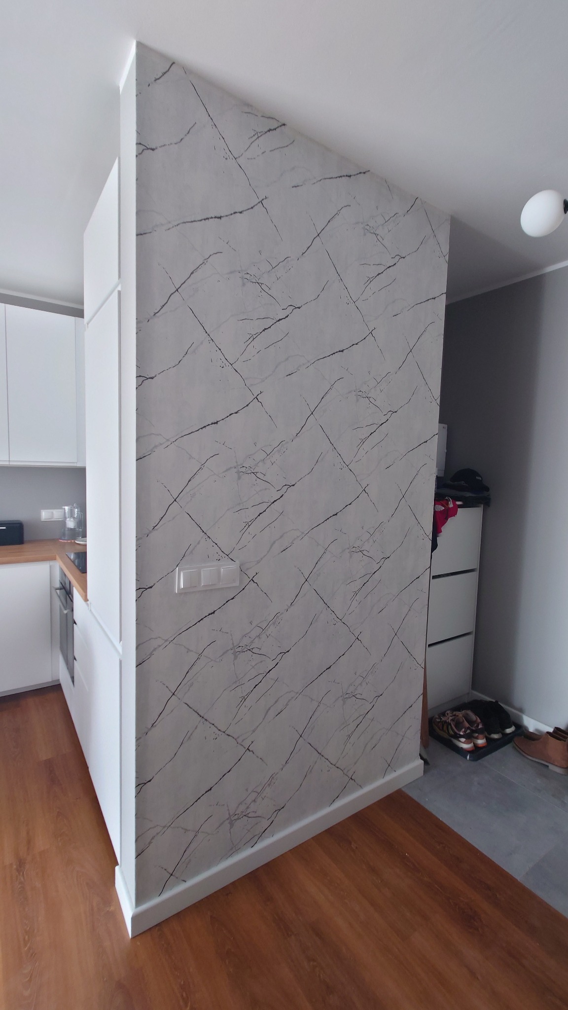 Ściana działowa w kuchni z tapetą imitującą marmur, widoczne gniazdka elektryczne, drewniana podłoga i fragment białej zabudowy kuchennej. Ukośna krawędź ściany.