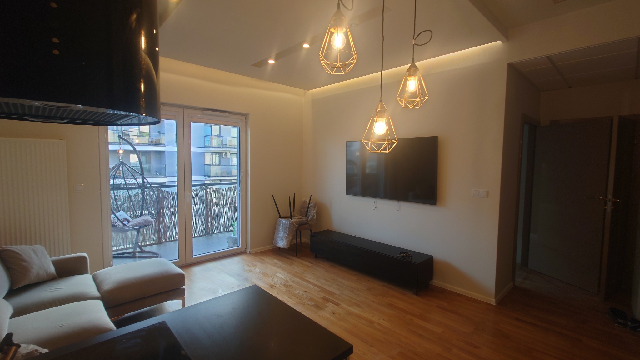 Jasne wnętrze salonu z drewnianą podłogą i balkonem. Widoczne lampy wiszące, telewizor na ścianie, sofa i krzesła zabezpieczone folią. Nowoczesny design.