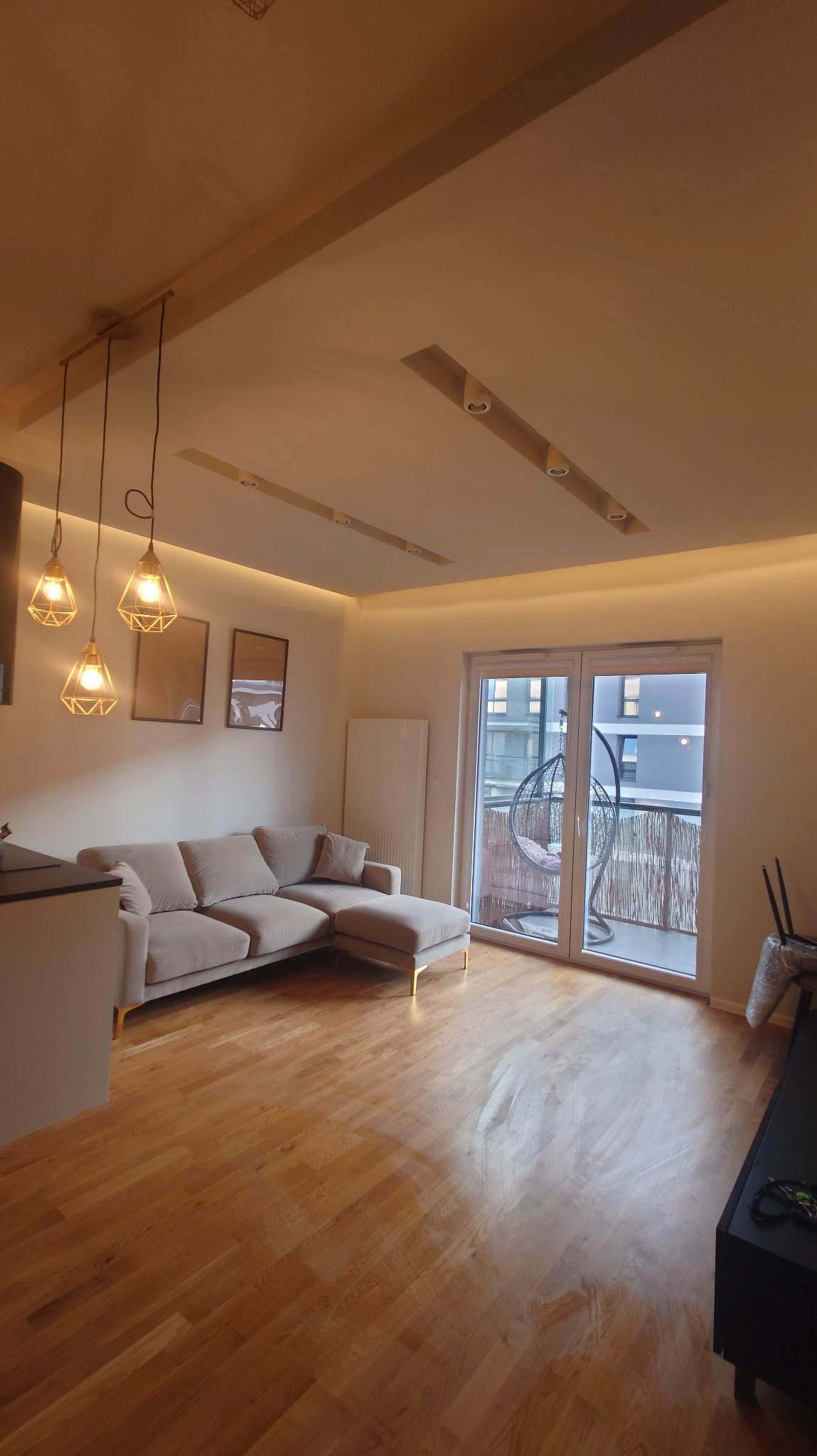 Jasny salon z drewnianą podłogą, beżową sofą i balkonem z huśtawką. Oświetlenie sufitowe LED i lampy wiszące dodają ciepła. Nowoczesne wnętrze.