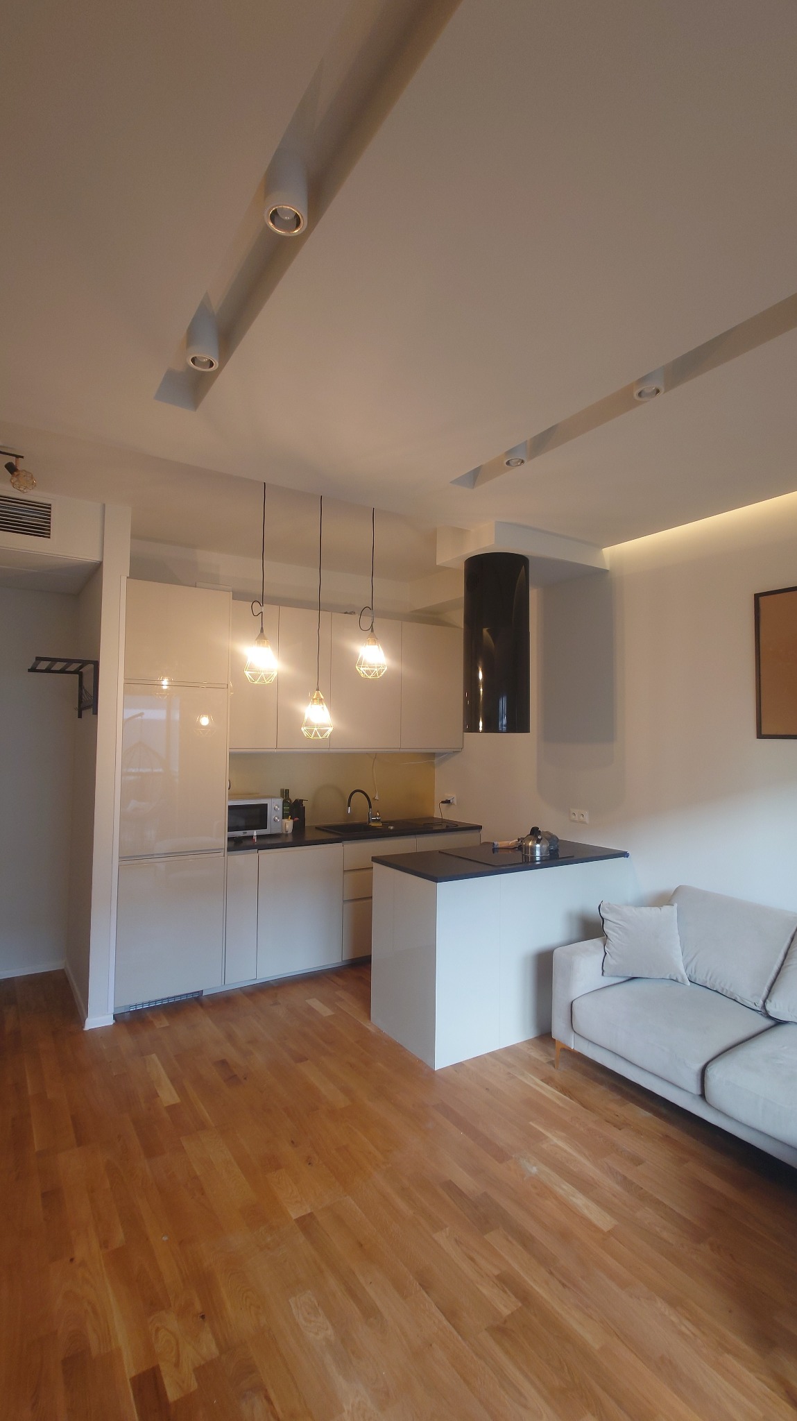 Jasne, nowoczesne wnętrze: drewniana podłoga, biała zabudowa kuchenna z wyspą i minimalistyczny salon. Oświetlenie halogenowe i wiszące lampy dodają ciepła.
