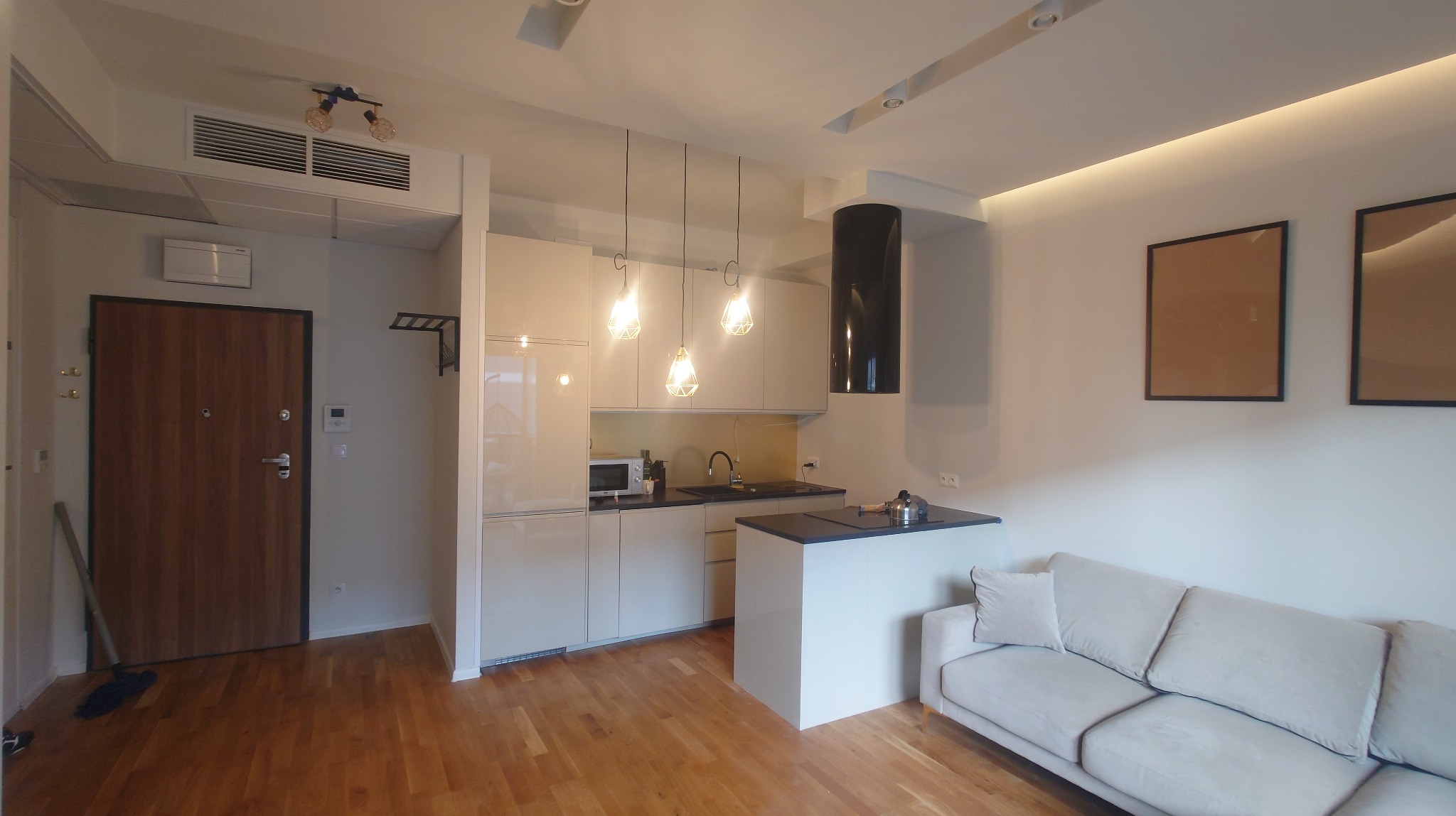 Nowoczesne wnętrze: parkiet, beżowa kuchnia z wyspą, sofa i minimalistyczne obrazy. Oświetlenie punktowe i wiszące lampy dodają elegancji.