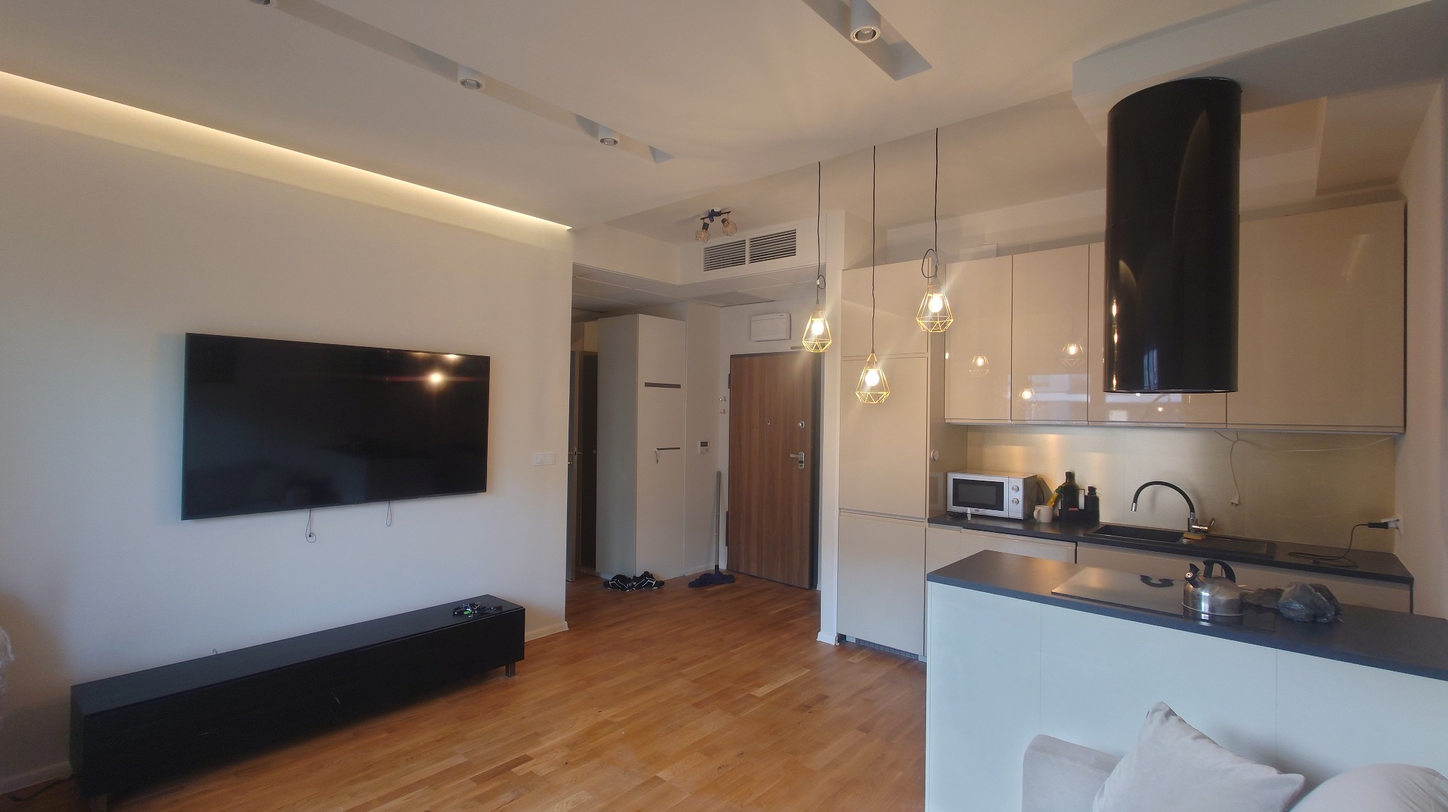 Jasne wnętrze mieszkania z drewnianą podłogą, widok na salon z telewizorem i otwartą kuchnią z nowoczesnym okapem oraz designerskimi lampami wiszącymi.