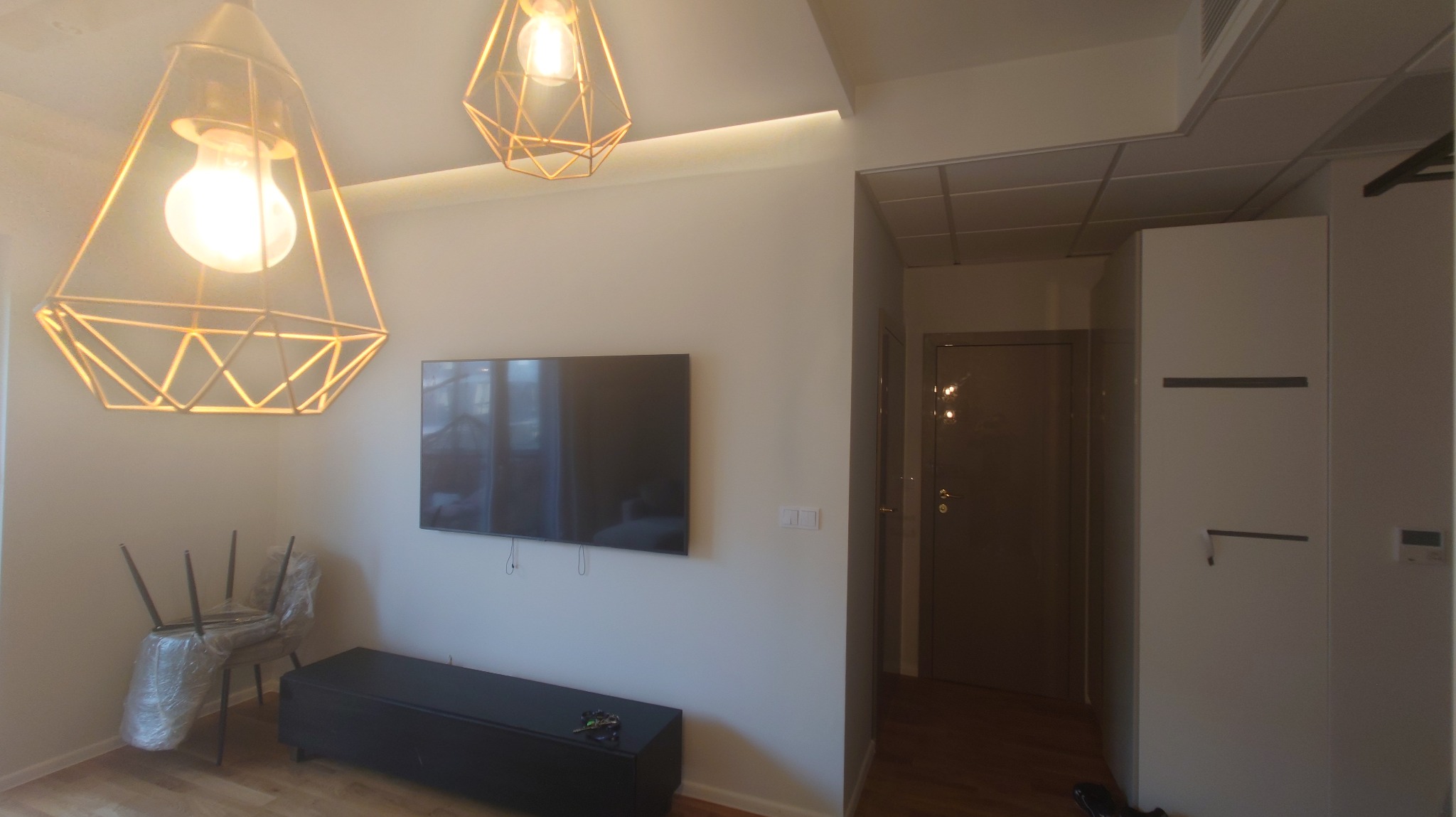 Jasne wnętrze po odświeżeniu: minimalistyczny salon z zawieszonym telewizorem, nowoczesnymi lampami i krzesłami zabezpieczonymi folią ochronną.