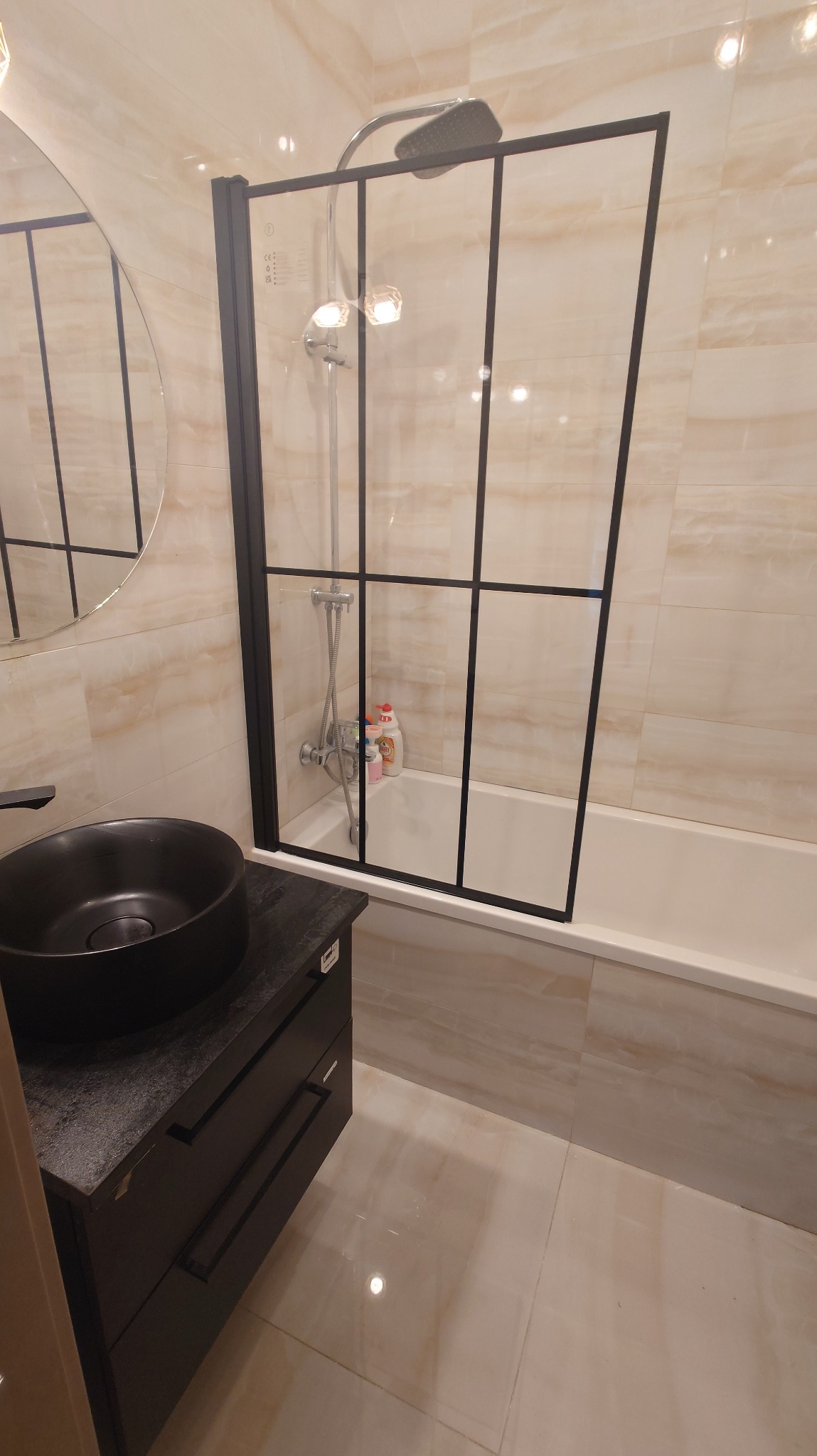 Nowoczesna łazienka z czarną umywalką nablatową, kabiną prysznicową typu walk-in z czarnymi szprosami i beżowymi płytkami imitującymi marmur.