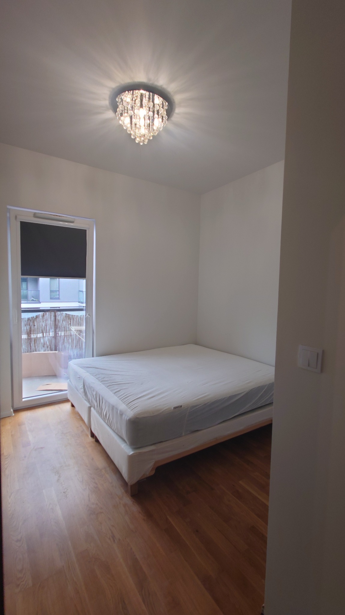 Jasne drewniane panele podłogowe w pokoju z balkonem i łóżkiem. Na suficie ozdobny żyrandol. Świeżo wykończone wnętrze.