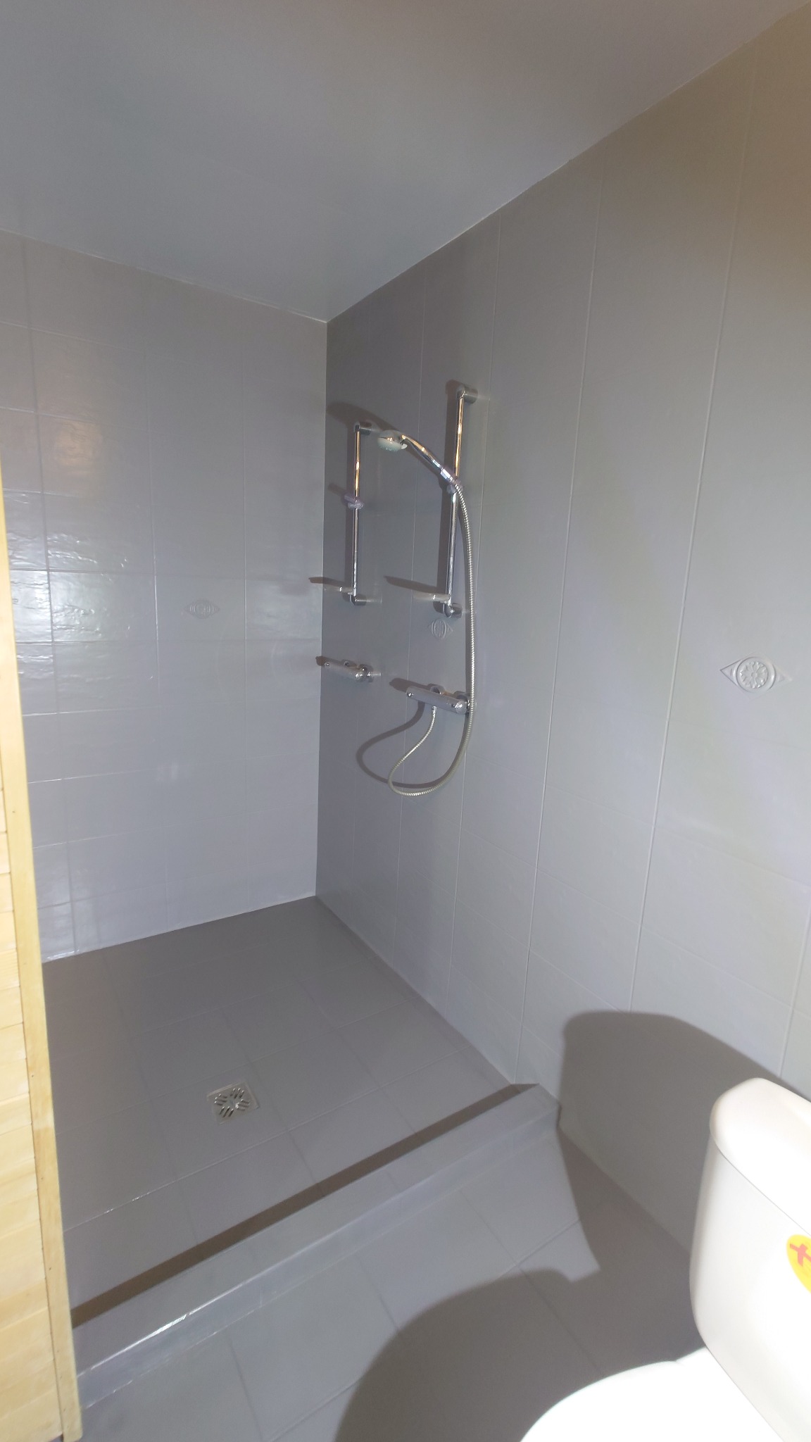 Szara, wykafelkowana kabina prysznicowa z chromowaną armaturą. Widoczny odpływ liniowy i fragment toalety. Minimalistyczny design, stonowana kolorystyka.