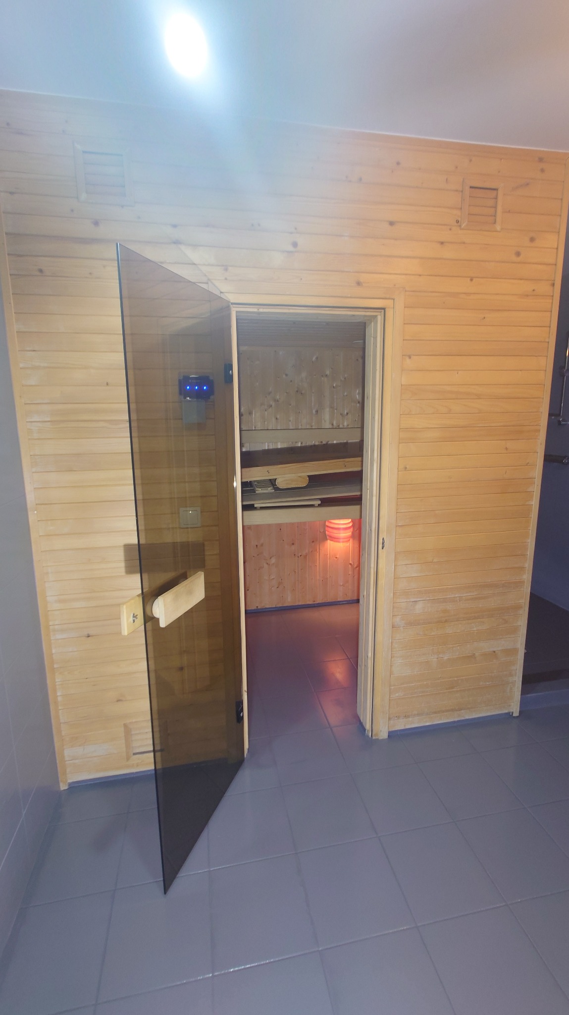 Jasna sauna z drewnianym wykończeniem, widoczna przez otwarte, szklane drzwi. Wnętrze oświetlone, z wyraźną strukturą drewna i widocznymi ławkami.