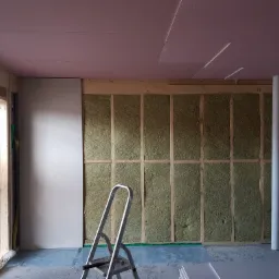 Wnętrze w trakcie prac wykończeniowych: ściana z izolacją z wełny mineralnej, częściowo obłożona płytami kartonowo-gipsowymi, widoczna drabina aluminiowa i różowa izolacja na suficie.