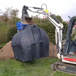 Mini koparka Bobcat podnosi czarny, plastikowy zbiornik z niebieską pokrywą nad wykopem w ziemi na trawniku. W tle drzewa i ogrodzenie.