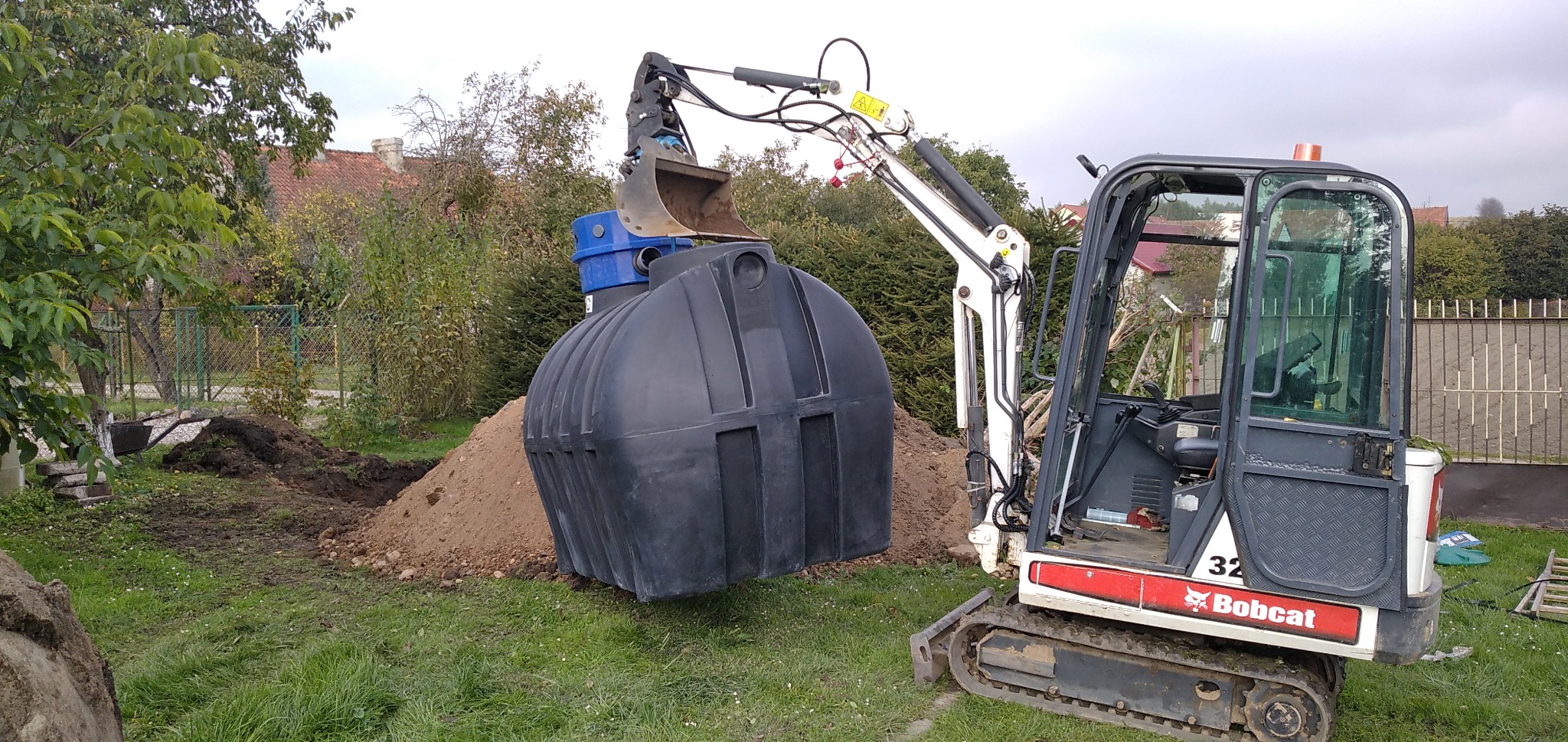 Mini koparka Bobcat podnosi czarny, plastikowy zbiornik z niebieską pokrywą nad wykopem w ziemi na trawniku. W tle drzewa i ogrodzenie.