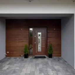 Nowoczesne wejście do domu z drewnianą okładziną ściany, szarą kostką brukową i białymi ścianami, widoczne doniczki z roślinami i częściowo zasłonięte okno z roletami.