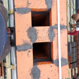 Wznoszenie komina z cegły klinkierowej na dachu, widoczna zaprawa murarska i miarka, dach pokryty membraną i łatami.