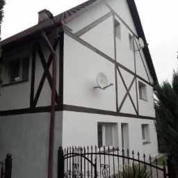 Dwukondygnacyjny dom z białą elewacją, brązowymi elementami dekoracyjnymi w stylu muru pruskiego i satelitami na ścianie, widziany zza czarnego, ozdobnego ogrodzenia.