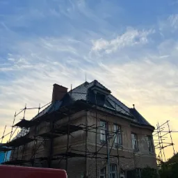 Kamienica w rusztowaniach z częściowo wymienionym dachem z ciemnej dachówki, widoczny fragment czerwonego autobusu na pierwszym planie, niebo z chmurami w tle.