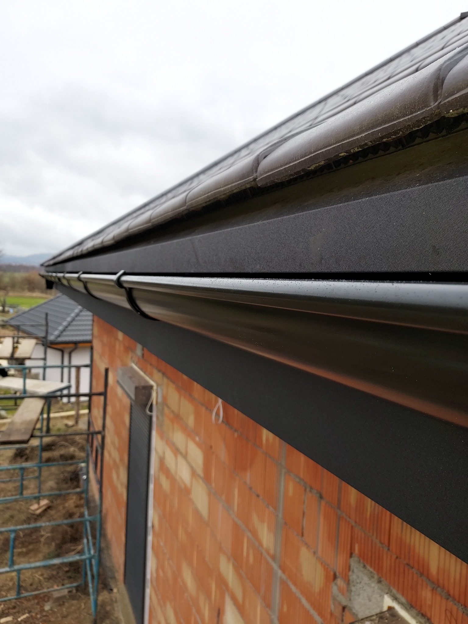 Nowo zamontowane, błyszczące, czarne rynny na tle ceglanej ściany budynku w trakcie budowy, widoczne rusztowania i fragment dachu sąsiedniego budynku.