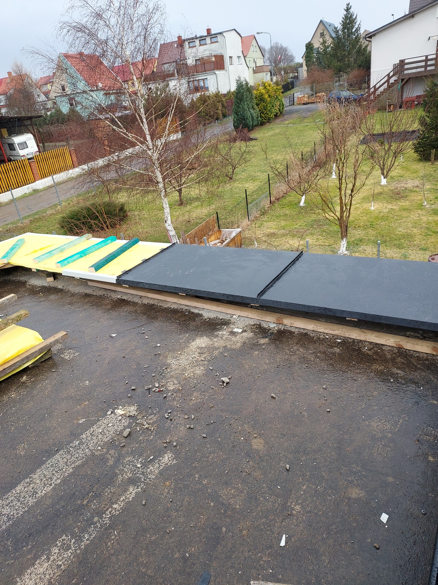 Fragment dachu w trakcie prac: żółta izolacja, zielone i białe elementy konstrukcyjne oraz czarne, płaskie pokrycie. W tle widok na okoliczne domy i drzewa.