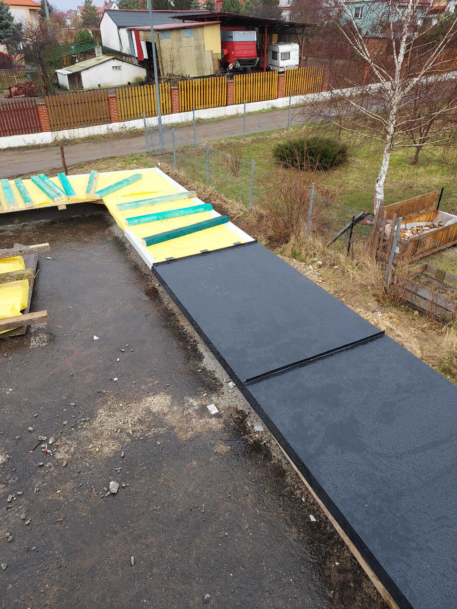 Widok z góry na fragment dachu w trakcie prac: czarne płyty izolacyjne na żółtej warstwie, zielone listwy, w tle ogród i zabudowania w pochmurny dzień.