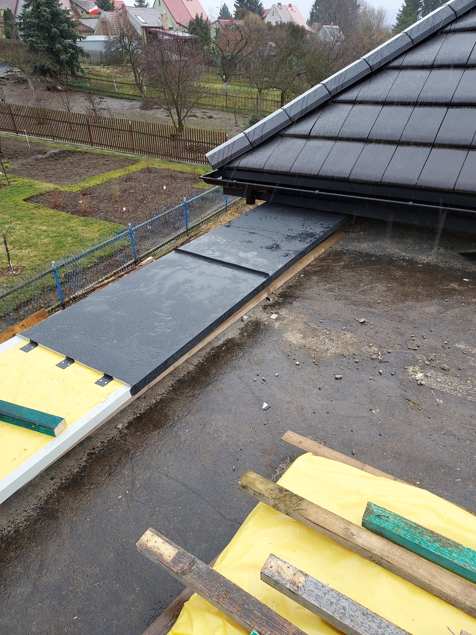 Wymiana dachu w trakcie: widoczne czarne płyty izolacyjne na dachu z szarymi dachówkami, żółta folia ochronna i drewniane elementy konstrukcyjne, deszczowa pogoda.