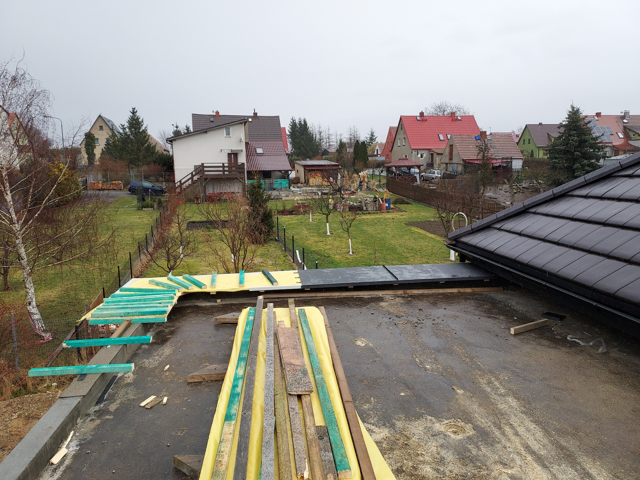 Widok z góry na płaski dach w trakcie prac: przygotowane drewniane elementy konstrukcyjne, żółta folia izolacyjna, częściowo ułożona czarna papa, w tle zabudowa jednorodzinna w pochmurny dzień.