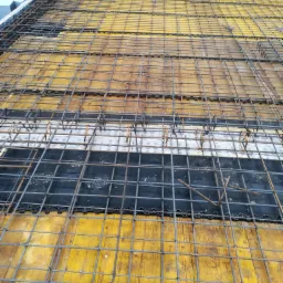 Zbrojenie płyty fundamentowej z siatki stalowej na żółtym szalunku, widoczne elementy izolacji i fragment zieleni w tle.